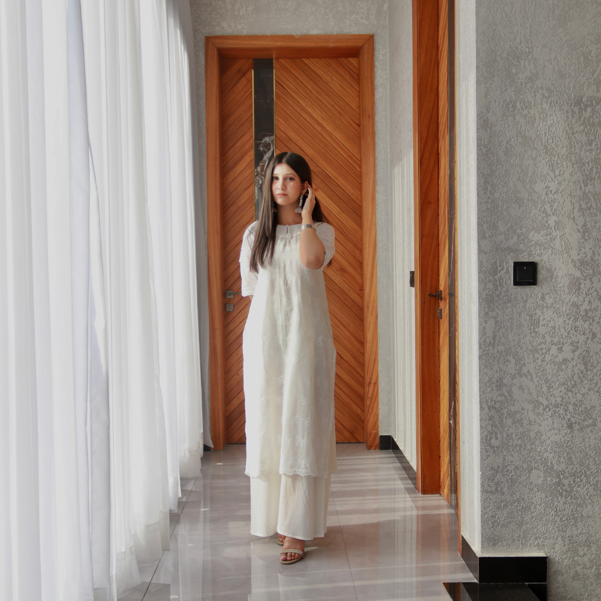 Woman in a white dress standing in a room with a wooden door and gray walls.